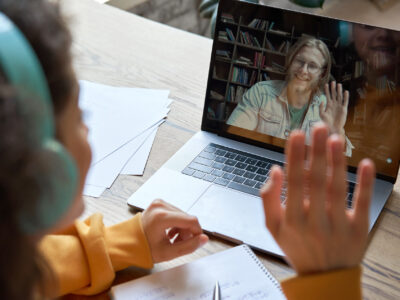 Hispanic teen girl school college student distance learning waving hand studying with online teacher on laptop screen. Elearning zoom video call, videoconference class with tutor. Over shoulder view.