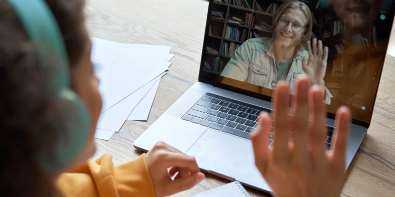 Hispanic teen girl school college student distance learning waving hand studying with online teacher on laptop screen. Elearning zoom video call, videoconference class with tutor. Over shoulder view.