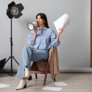 Young happy female film director with megaphone, script and studio lighting sitting on chair against grey background