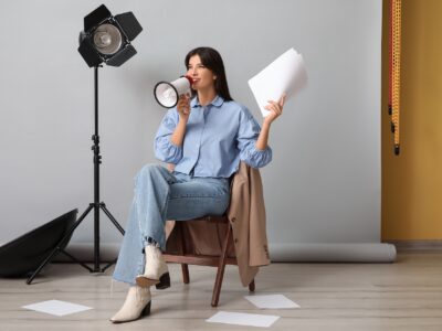 Young happy female film director with megaphone, script and studio lighting sitting on chair against grey background