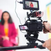 Vibrant Scene Captures Young Asian Female Host In Pink Blazer On Film Set, Engaging In Lively Discussion, With Focus On Professional Video Equipment And Dynamic Set Design.