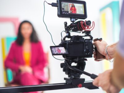 Vibrant Scene Captures Young Asian Female Host In Pink Blazer On Film Set, Engaging In Lively Discussion, With Focus On Professional Video Equipment And Dynamic Set Design.
