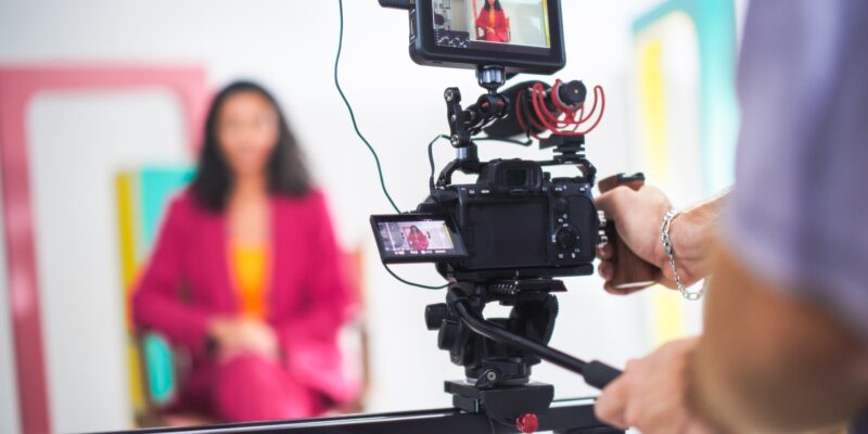 Vibrant Scene Captures Young Asian Female Host In Pink Blazer On Film Set, Engaging In Lively Discussion, With Focus On Professional Video Equipment And Dynamic Set Design.