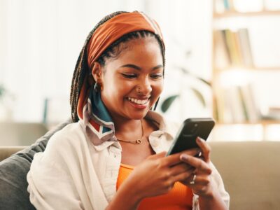 Smile, online and woman with smartphone in home for social media, reading forum or ebook. African person, mobile and relax on sofa with chat app for communication, text notification or download game