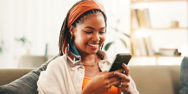 Smile, online and woman with smartphone in home for social media, reading forum or ebook. African person, mobile and relax on sofa with chat app for communication, text notification or download game