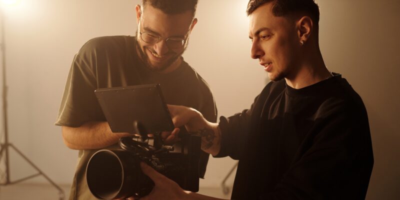 Waist up shot of two young male videographers reviewing footage on camera monitor during music video shoot in studio