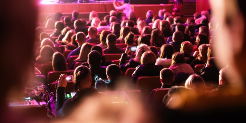 Spectators sit in the hall and watch a concert. People in the auditorium watching the performance. Theater audience. Music concert or festival. Out of focus.