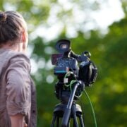 Rear view to man videographer recording outdoor movie about nature, back view to TV channel male crew with TV camera, green background. Long haired man with professional camera records video for TV