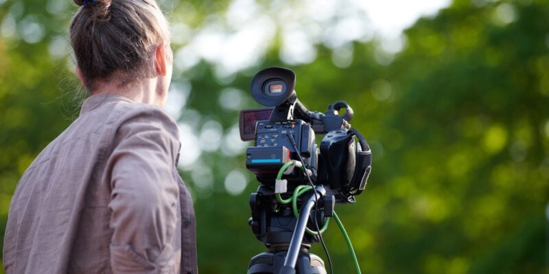 Rear view to man videographer recording outdoor movie about nature, back view to TV channel male crew with TV camera, green background. Long haired man with professional camera records video for TV