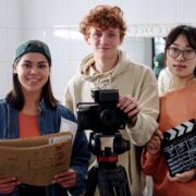 Portrait of three young people holding film equipment and smiling warmly at camera in studio setting while standing near large mirror in background