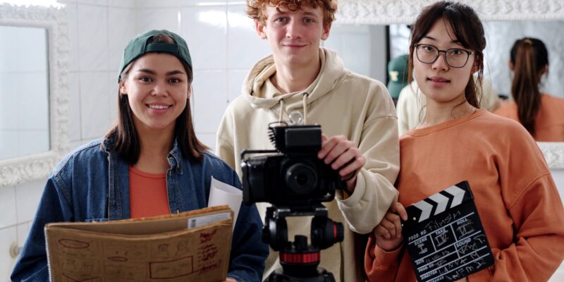 Portrait of three young people holding film equipment and smiling warmly at camera in studio setting while standing near large mirror in background